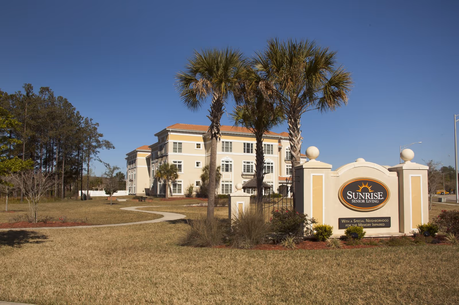 Front exterior of the Sunrise Senior Living building with palm trees and a large entrance sign on a sunny day.
