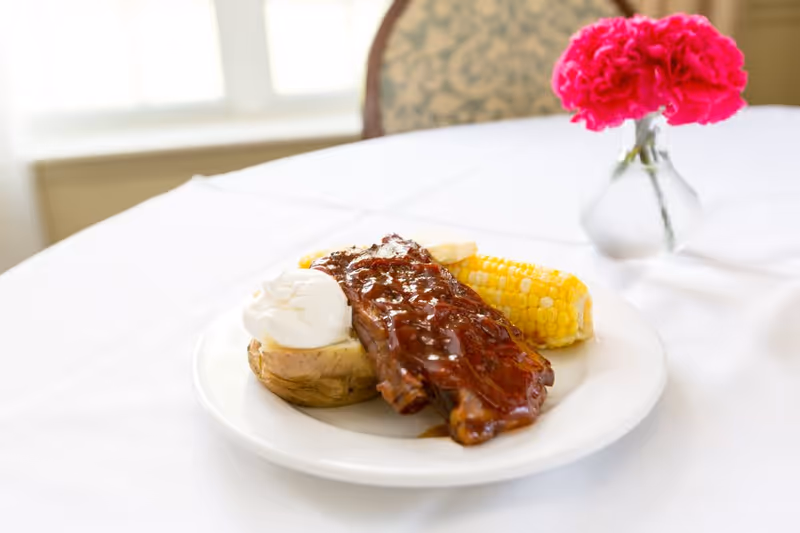 A white plate on a white tablecloth holding a serving of barbecue ribs, a baked potato topped with sour cream, and a piece of corn on the cob. In the background, there is a small vase with bright pink flowers and a chair with a patterned backrest.