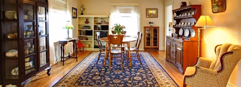 Bright traditional dining room with a wooden table and chairs centered on a patterned rug, flanked by china cabinets and an upholstered armchair.