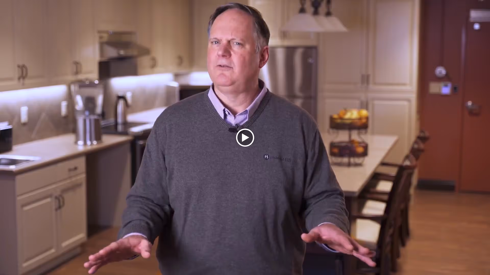 A man wearing a gray sweater stands in a well-lit kitchen with cream-colored cabinets, a stainless steel refrigerator, and a long island with chairs. The kitchen has modern appliances and a bowl of fruit on the counter.