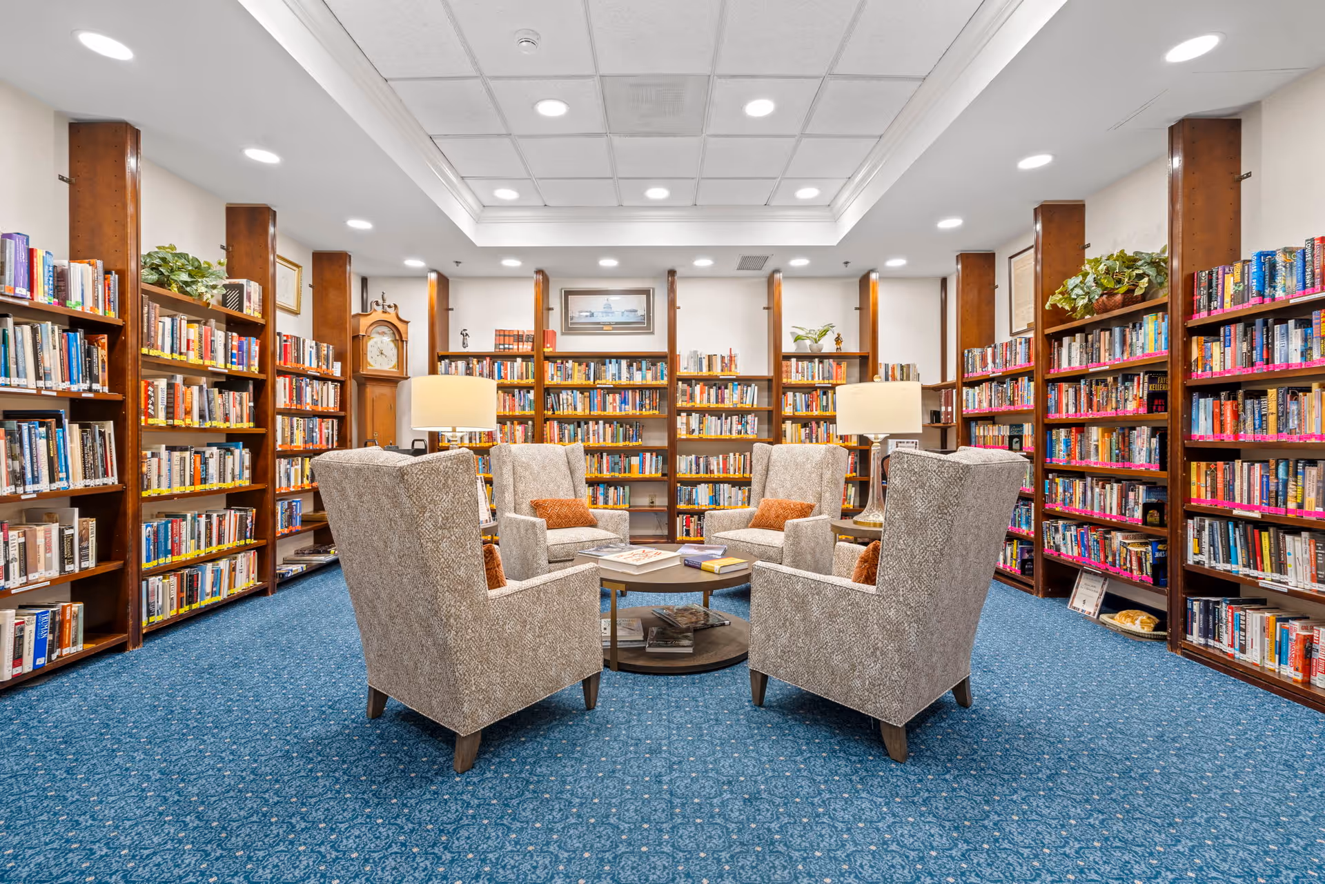 A cozy library room with blue carpet and wooden bookshelves filled with books lining the walls. Four beige upholstered armchairs with orange cushions are arranged around a circular coffee table with books on it. Two table lamps provide warm lighting, and a grandfather clock is visible in the corner. The ceiling has recessed lighting and a white tray ceiling design.