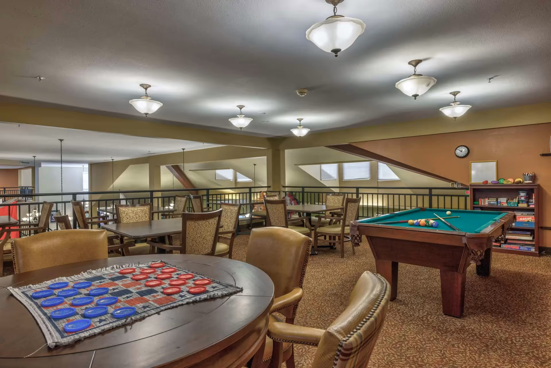 A spacious recreational room with a pool table and a round table set up for checkers. The room has multiple tables and chairs, carpeted floor, warm lighting from ceiling fixtures, and a bookshelf filled with board games and books against the wall.