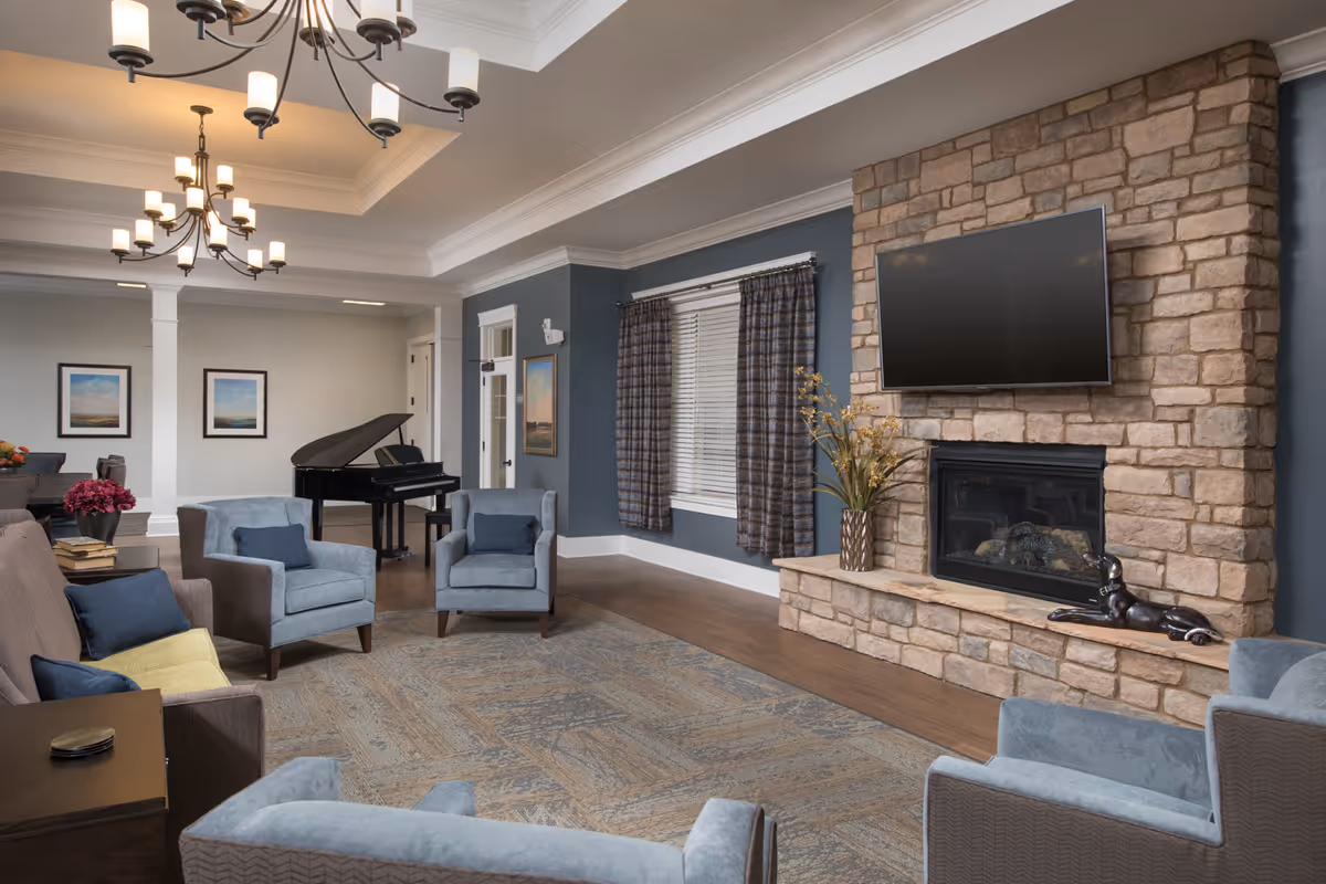 A cozy senior living room with a stone fireplace, a mounted flat-screen TV above it, and a decorative sculpture on the hearth. The room features blue-gray walls, a large area rug, multiple comfortable armchairs and a sofa with blue cushions, a grand piano in the background, and framed artwork on the walls. Two chandeliers hang from the ceiling, and a window with plaid curtains is visible.