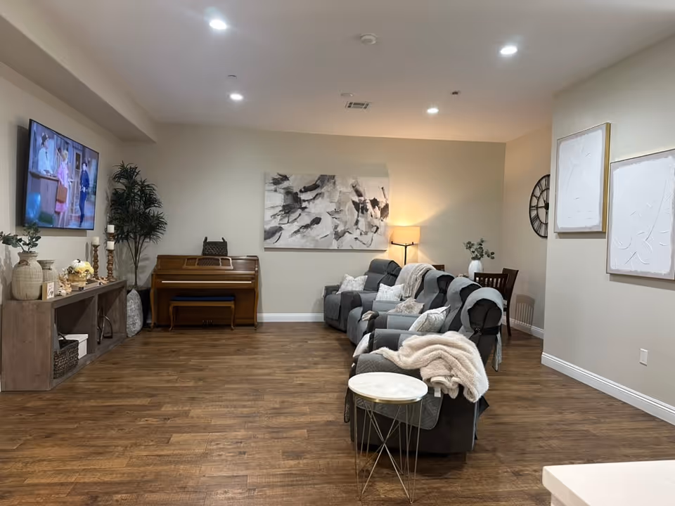 A cozy senior living common area with a row of gray reclining chairs covered with blankets and pillows, a wooden piano against the back wall, a flat-screen TV mounted on the left wall, decorative plants, and abstract artwork on the walls. The room has wooden flooring and soft lighting from ceiling lights and a floor lamp.