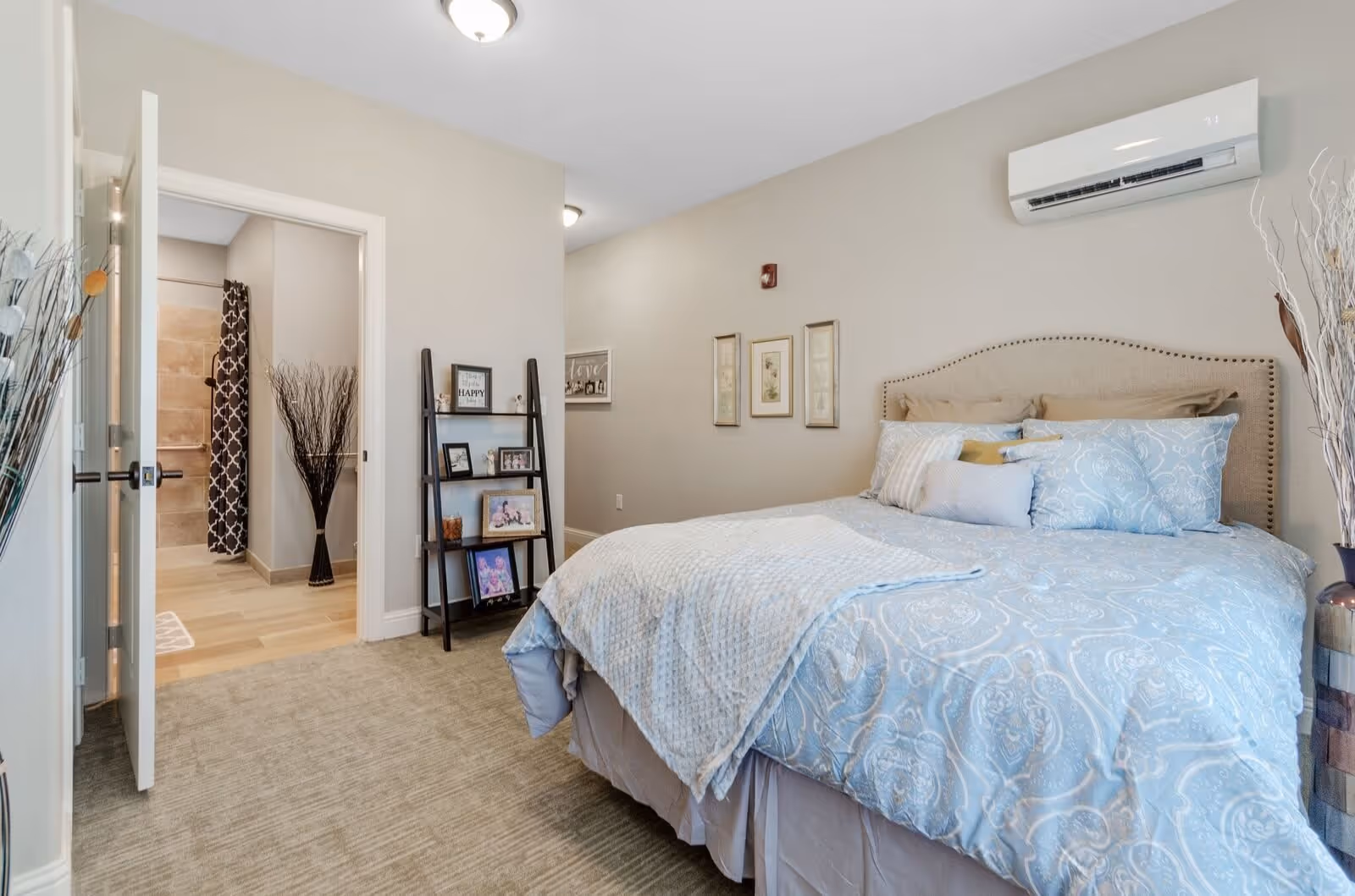 A cozy bedroom with a large bed covered in light blue patterned bedding and multiple pillows. The room has beige walls with three framed pictures hanging above the bed. There is a black ladder-style shelf with framed photos and decor items near the open door leading to a bathroom. The bathroom has beige tiles and a dark patterned shower curtain. Decorative tall vases with dried branches are placed in the bedroom corners.