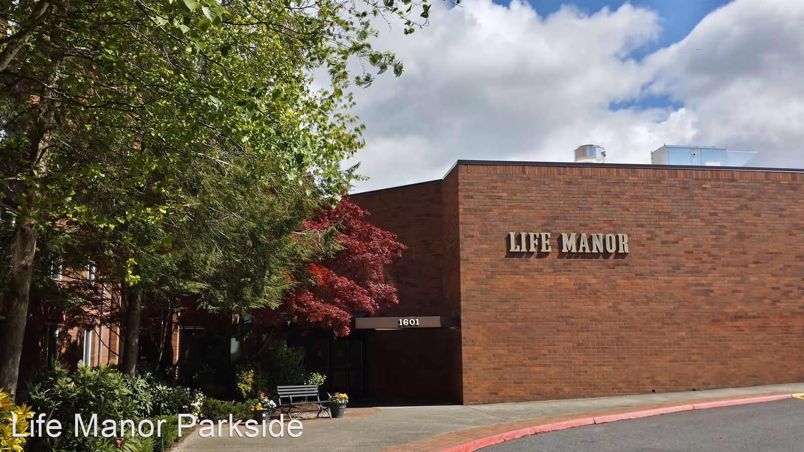 Front exterior of a brick building labeled "Life Manor" with an entrance, trees, and a bench.