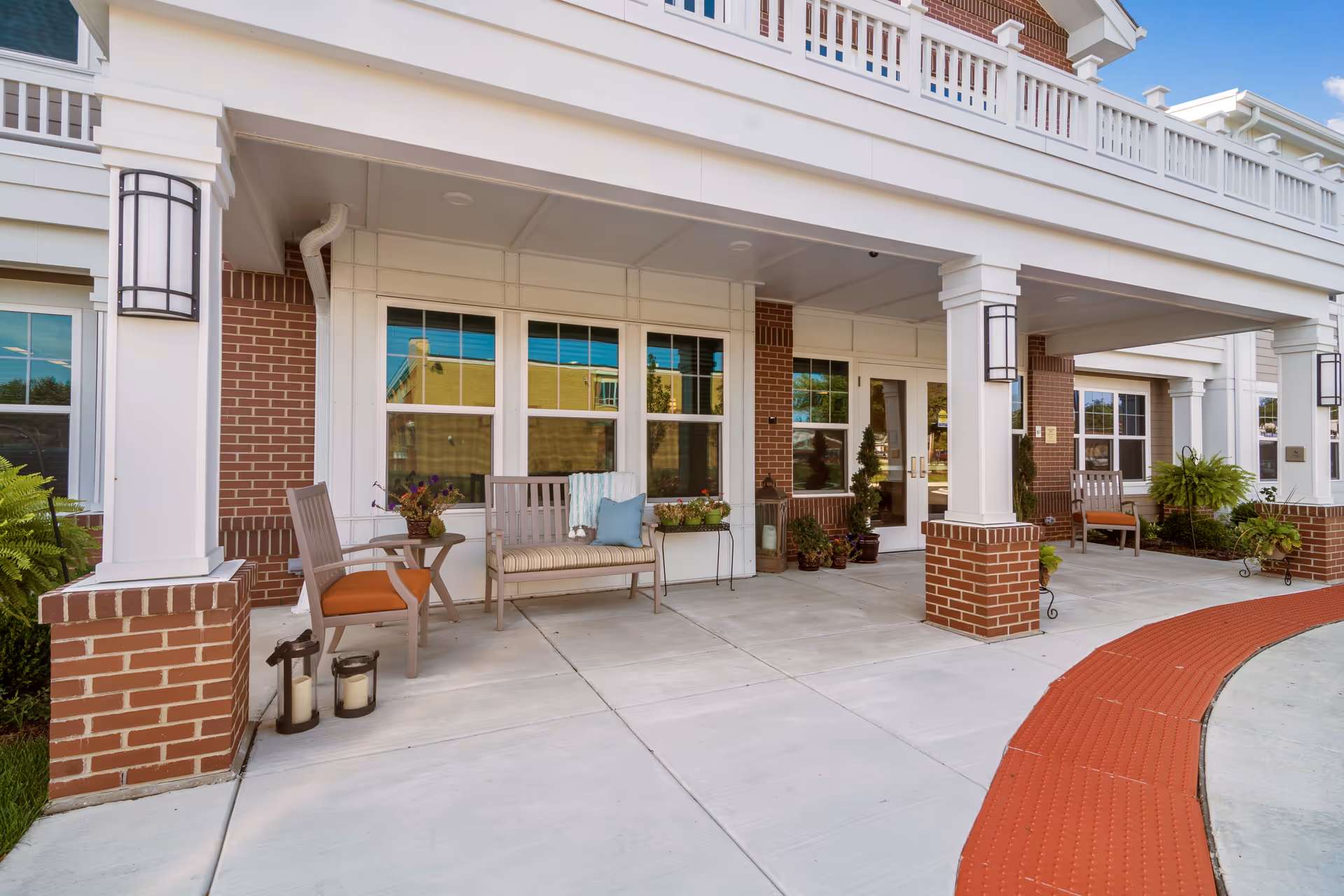 Covered outdoor seating area at the entrance of a building with brick pillars and white trim. The area features chairs, a bench with cushions, small tables, potted plants, and lanterns. The building has large windows and double glass doors, with a red textured walkway curving in front.