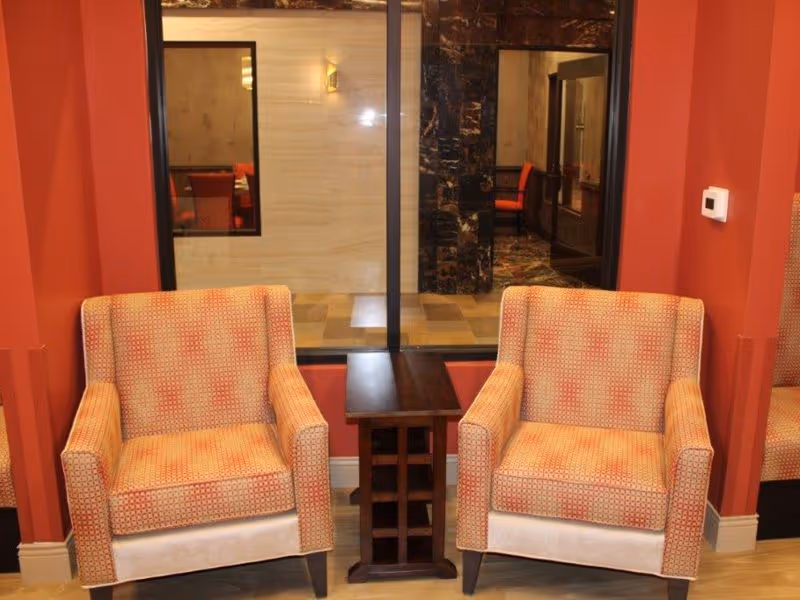Two patterned armchairs flanking a small wooden side table in a red-walled interior seating area with a window into a hallway.
