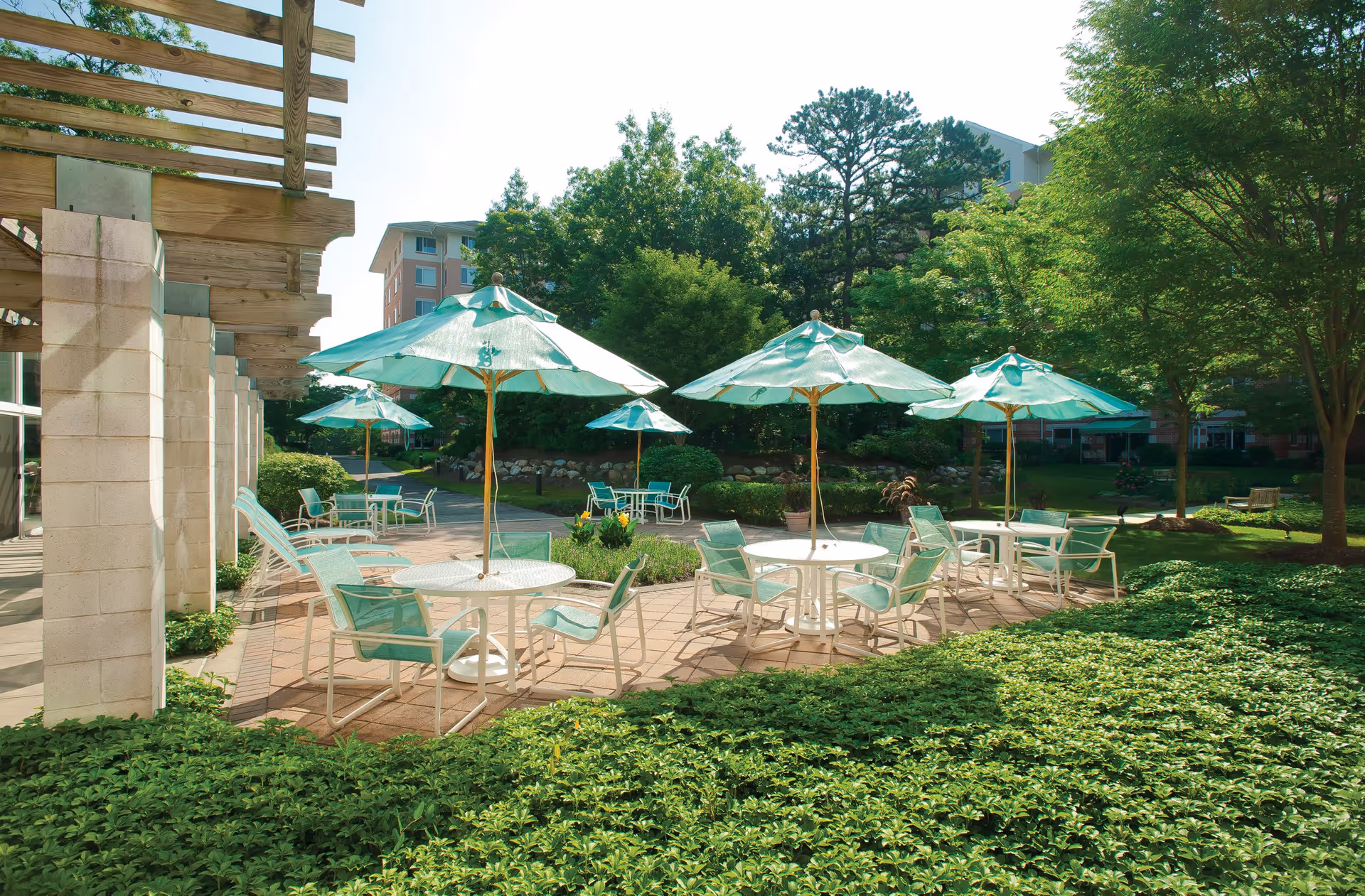 Outdoor patio area with several round tables and chairs, each table shaded by a teal umbrella. The patio is surrounded by green bushes and trees, with a building visible in the background under a clear sky.