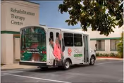 Entrance of a Rehabilitation Therapy Center with a white shuttle bus parked in front displaying an advertisement featuring smiling older adults.