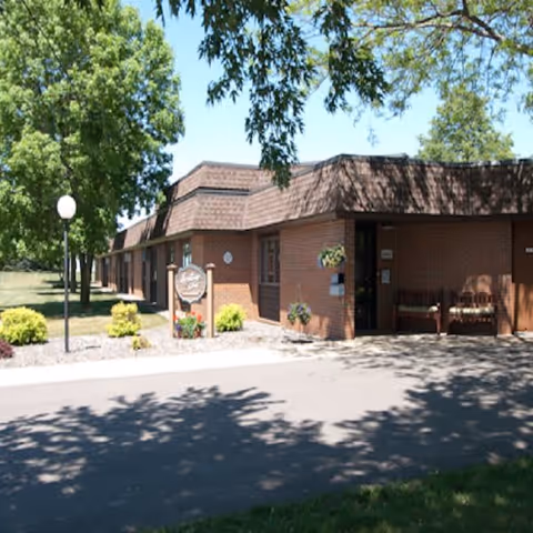 Exterior view of a single-story brick building with a brown roof, surrounded by trees and landscaping. There is a paved driveway in front and a sign near the entrance that reads Heritage Court. Two benches are placed near the entrance under a shaded area.