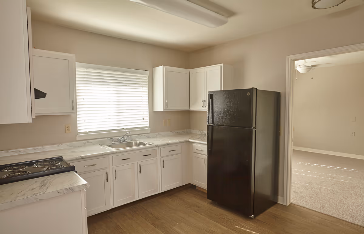 A kitchen with white cabinets, marble-patterned countertops, a black refrigerator, and a stove. There is a window with white blinds above the sink, and an adjacent room with carpet and a ceiling fan is visible through a doorway.