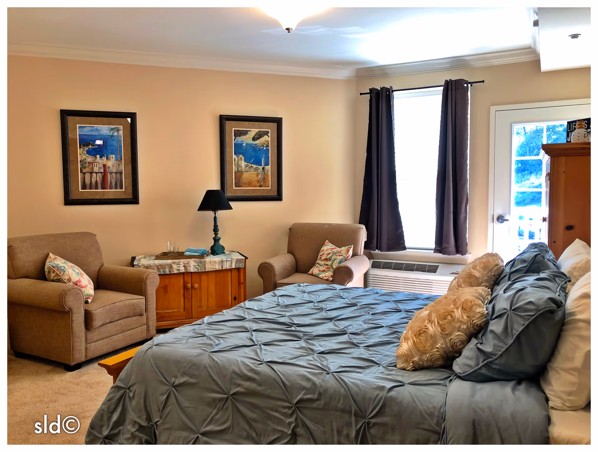 Well-lit bedroom with a made bed, two armchairs, a nightstand with lamp, framed artwork, and a curtained window.