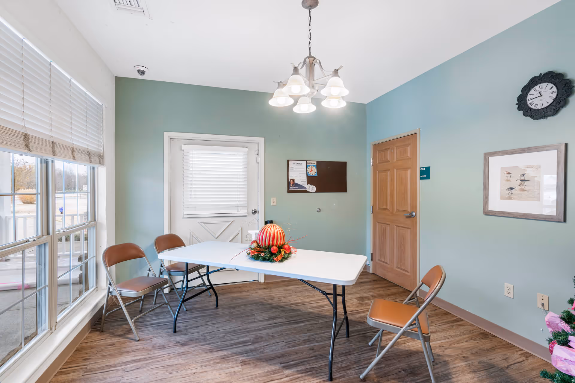 A small room with light green walls and wood flooring featuring a white folding table with a festive centerpiece and four brown folding chairs around it. There is a door with a window covered by blinds, a bulletin board on the wall, a clock, and a framed picture of birds. Large windows with blinds let in natural light.