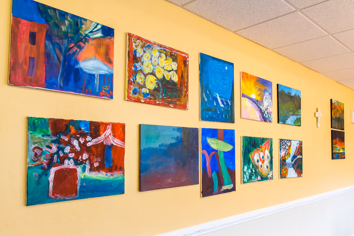 A yellow wall decorated with multiple colorful abstract paintings arranged in two rows, with a white chair rail molding below and a white ceiling with tiles above.