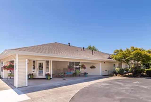 Exterior view of a single-story building with a light-colored roof and siding, featuring a covered entrance area with chairs and potted plants. The building is surrounded by a paved driveway and some greenery, including trees and shrubs, under a clear blue sky.