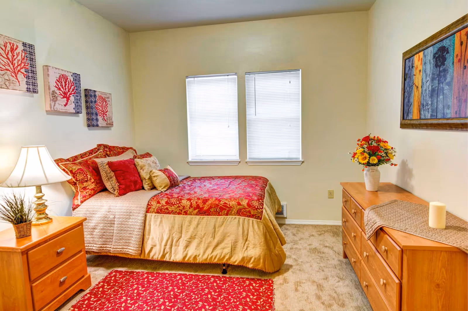 A cozy bedroom with a bed covered in red and gold bedding, multiple decorative pillows, a wooden nightstand with a lamp and a small plant, two windows with closed blinds, a wooden dresser with a vase of flowers and a candle, and framed artwork on the walls.