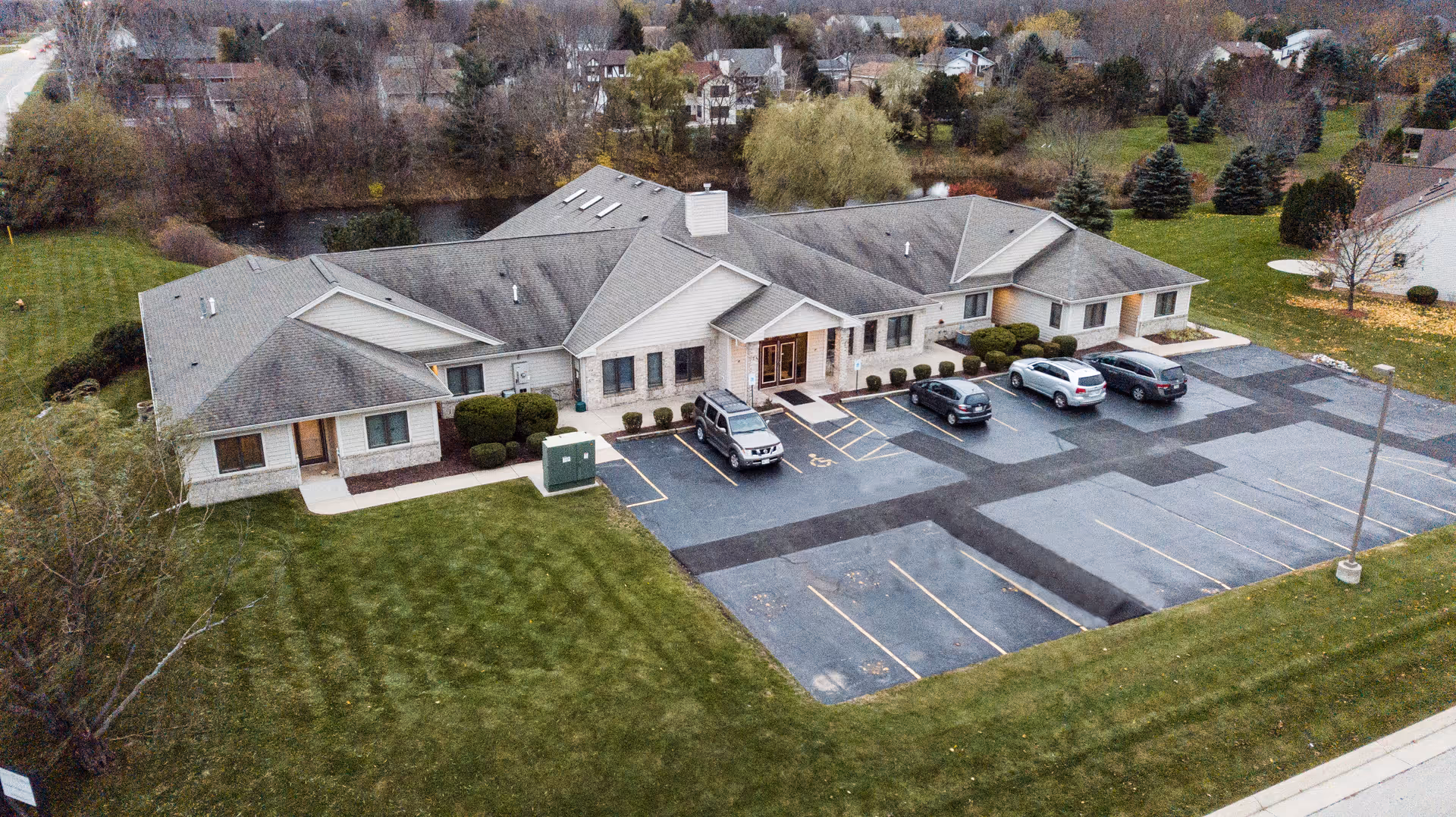 Aerial view of a single-story assisted living facility building with a gray roof and beige exterior walls. The building is surrounded by a well-maintained lawn and a parking lot with several parked cars. Trees and residential houses are visible in the background.