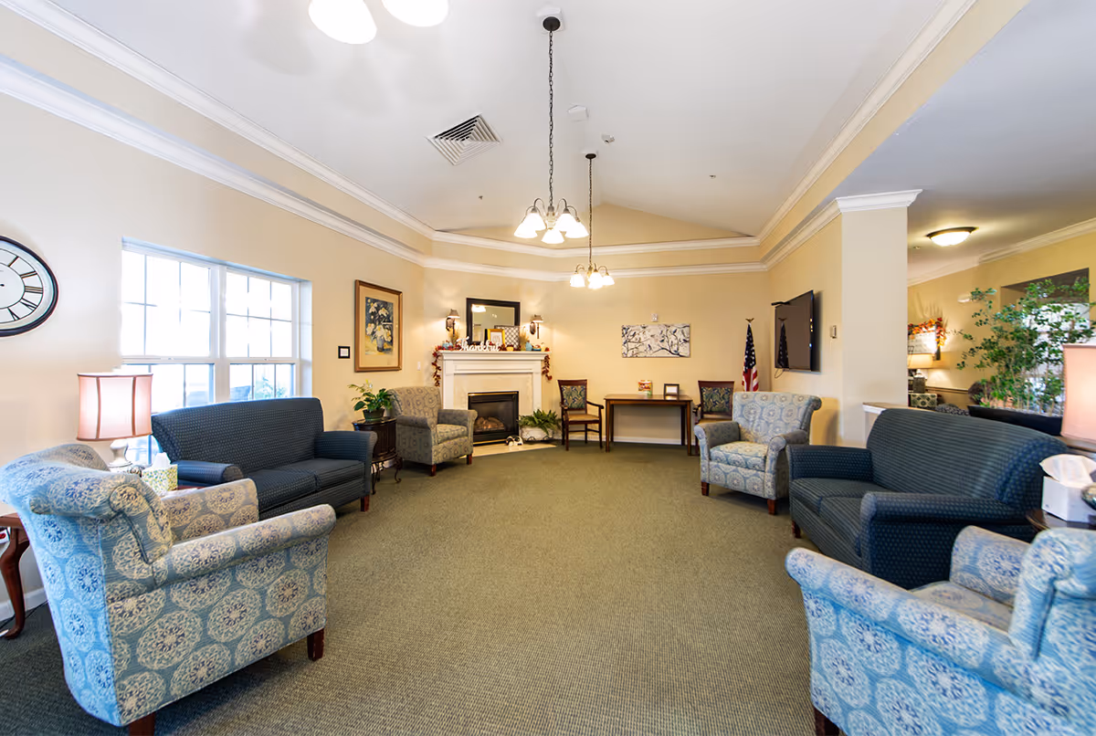 Bright communal lounge with upholstered armchairs and sofas arranged around a fireplace and TV.