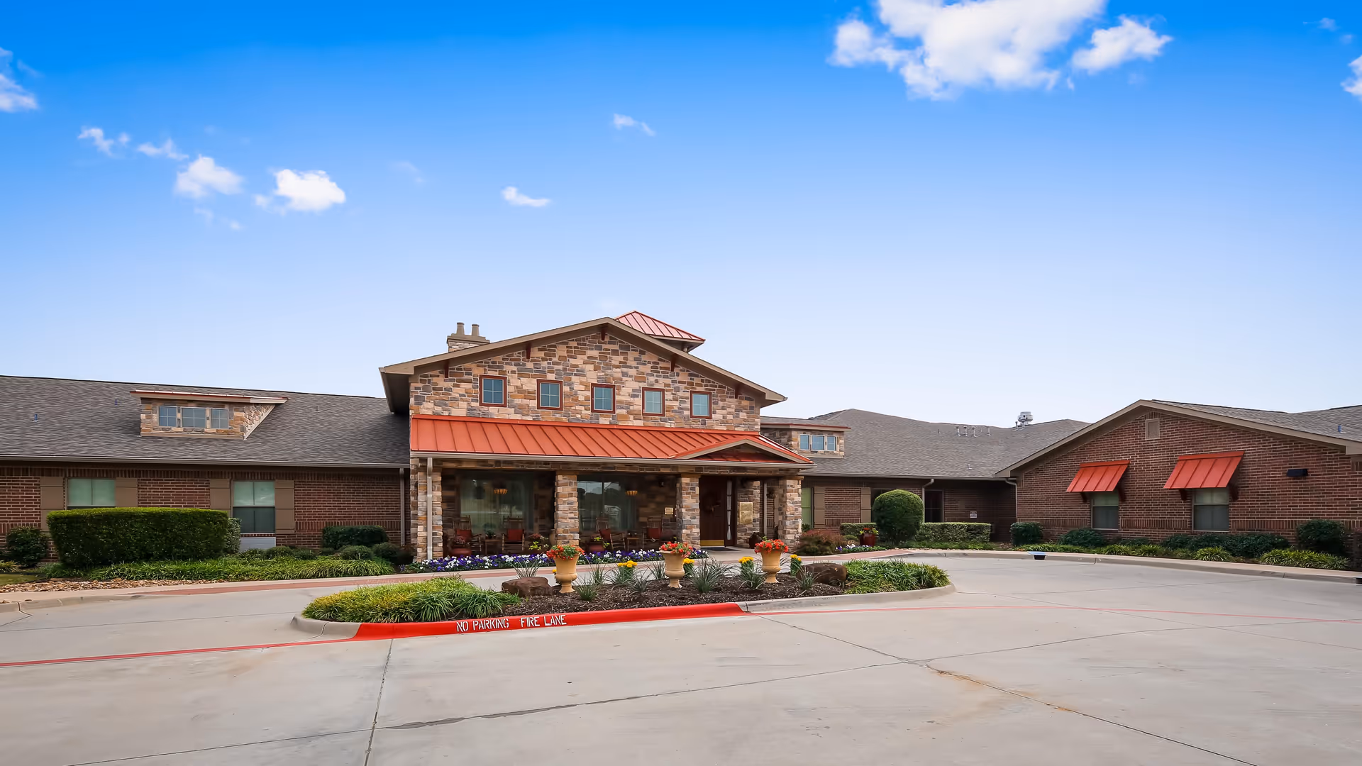 Front exterior view of Willow Bend Assisted Living & Memory Care facility with a stone and brick facade, a covered entrance with a red metal roof, landscaped flower beds, and a clear blue sky.