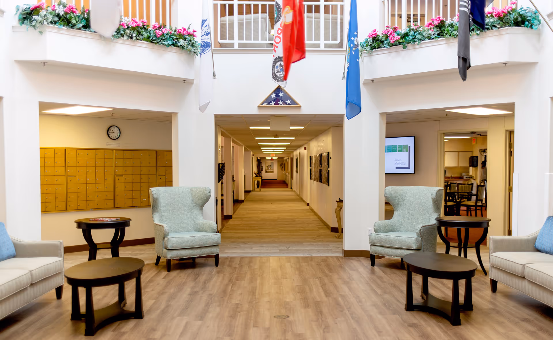Bright senior living facility lobby with armchairs and small tables facing a long hallway, flags overhead and floral planters on the upper balcony.