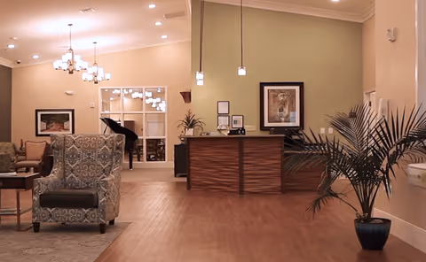 A spacious and well-lit common area with wooden flooring, featuring a patterned armchair, a small table, a reception desk with framed artwork on the wall behind it, a large potted plant, and a grand piano visible through glass doors in the background.