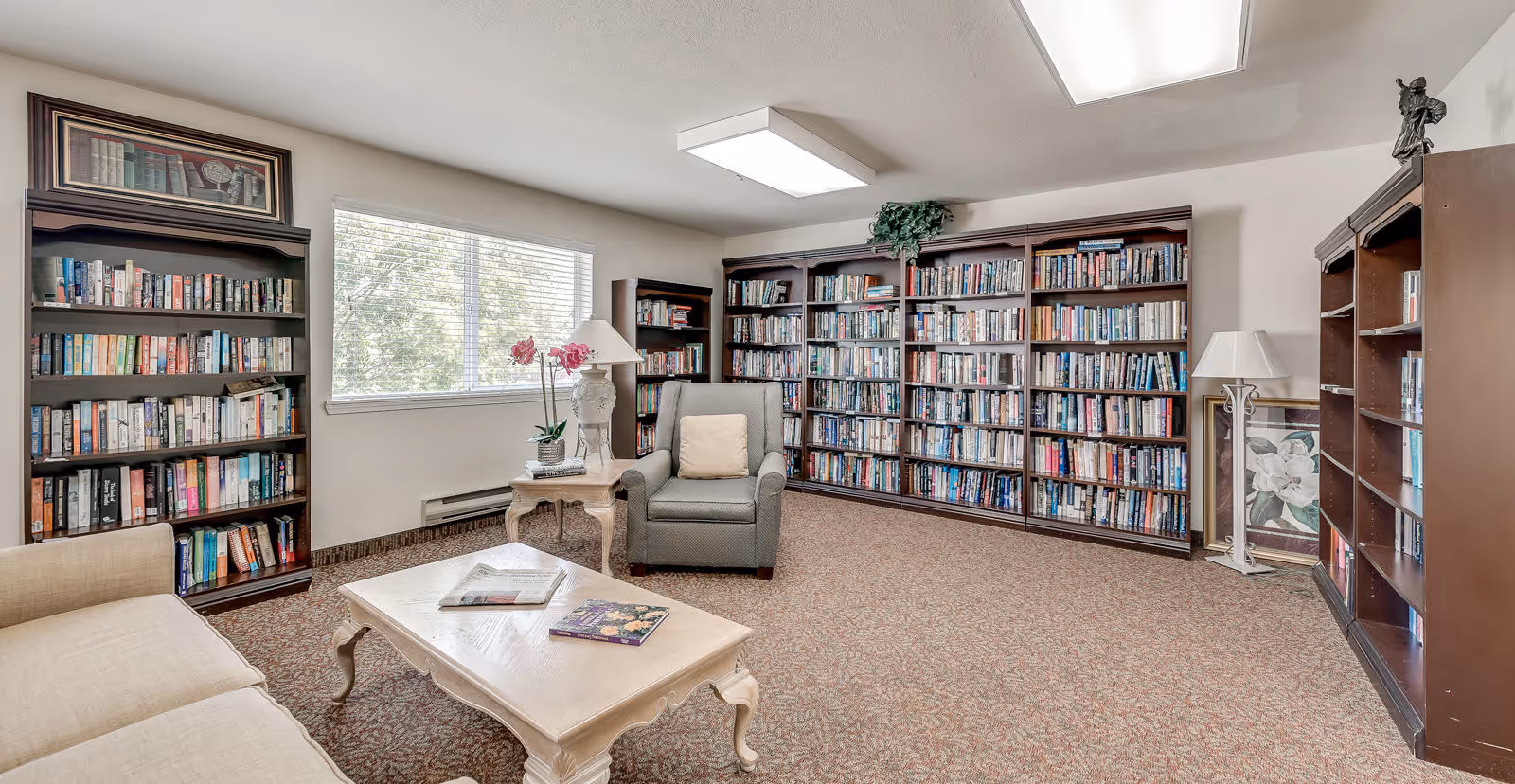 A cozy reading room with multiple dark wood bookshelves filled with books along the walls. There is a large window letting in natural light, a beige sofa, a gray armchair with a cushion, a white coffee table with magazines, and a side table with a lamp and a pink orchid. The room has a carpeted floor and a bright ceiling light.