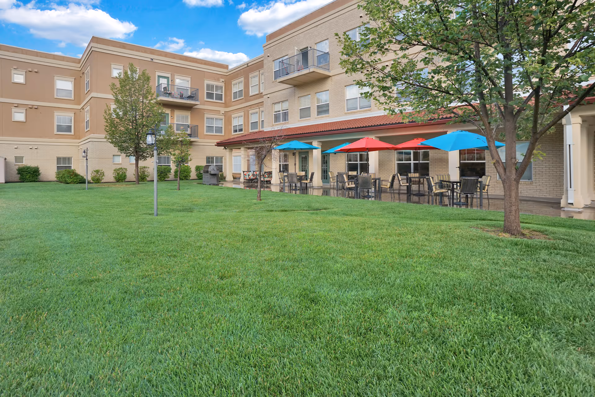 Outdoor view of The Inn at Greenwood Village showing a well-maintained green lawn with several trees and a patio area with tables and chairs shaded by colorful umbrellas in red and blue. The building has multiple floors with balconies and large windows under a partly cloudy sky.