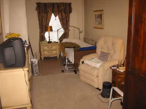 Senior living facility bedroom with bed, recliner, dresser, TV, and window curtains.