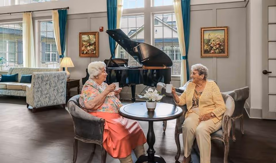 Two elderly women sit at a small table drinking coffee in a bright lounge with a grand piano and sofas behind them.