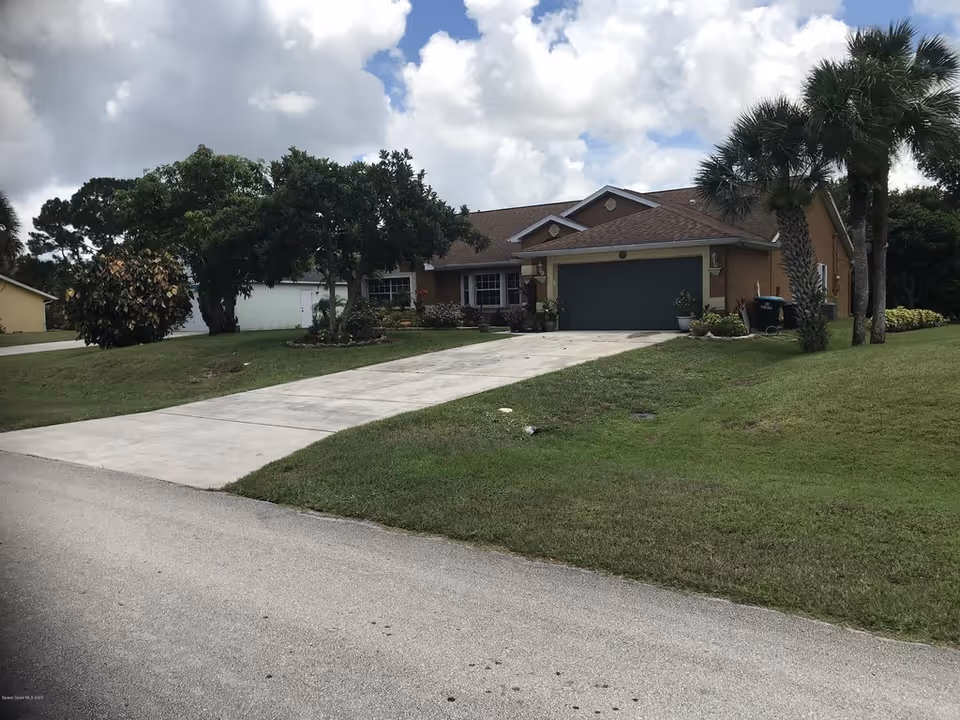 Single-story house with a two-car garage, concrete driveway, front lawn and palm trees under a cloudy sky.