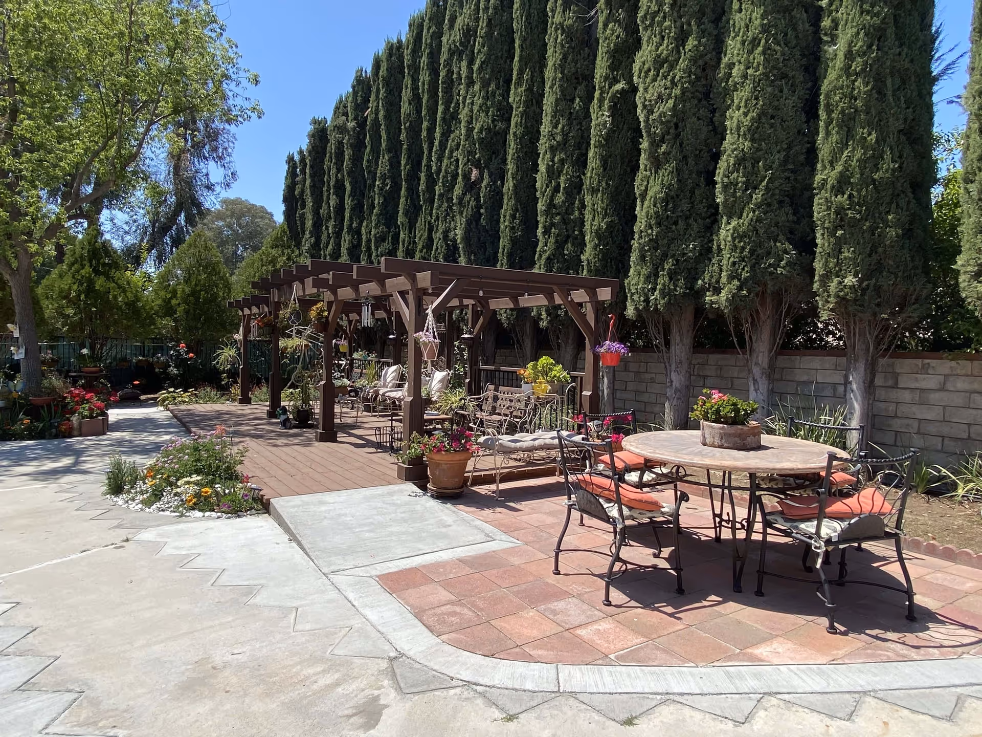 Outdoor patio area with a round table and four chairs with cushions, surrounded by tall trees and various potted plants. There is a wooden pergola with seating underneath and hanging plants, set against a clear blue sky.