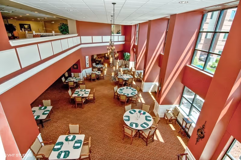 Two-story dining room with round and square tables set with green place mats, tall windows, and a central chandelier.
