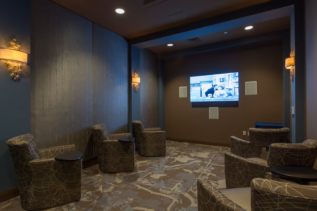 A cozy media room with six patterned armchairs arranged in two rows facing a wall-mounted flat-screen TV playing a black and white show. The room has dim lighting with decorative wall sconces and a patterned carpet.