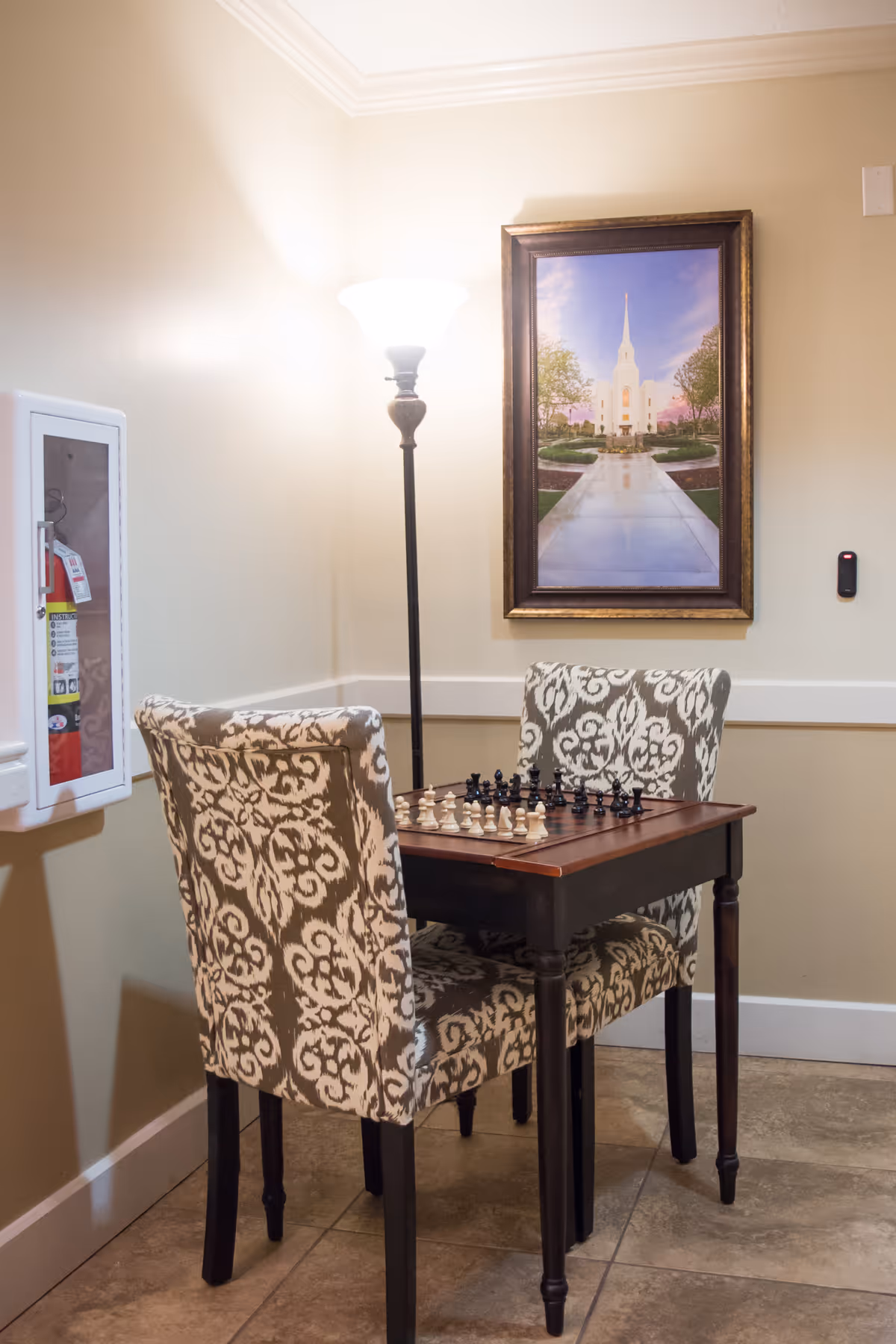 A small seating area with two patterned upholstered chairs and a wooden table set up for a chess game. A floor lamp is positioned behind the table, and a framed picture of a white building with a tall spire hangs on the beige wall. A fire extinguisher cabinet is mounted on the left wall.