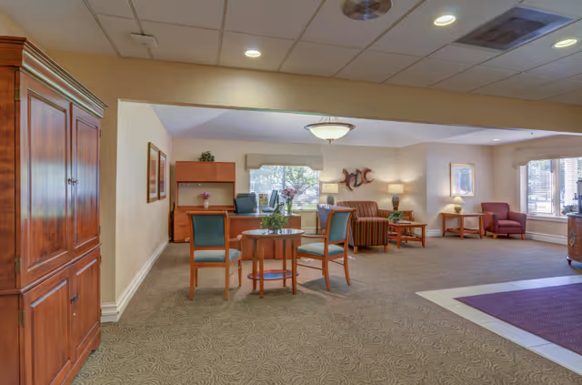 A spacious, well-lit living room area in a senior living facility featuring a round wooden table with four chairs, a wooden cabinet on the left, a desk with a chair and computer in the background, and a seating area with a striped sofa, armchairs, and side tables with lamps near large windows.
