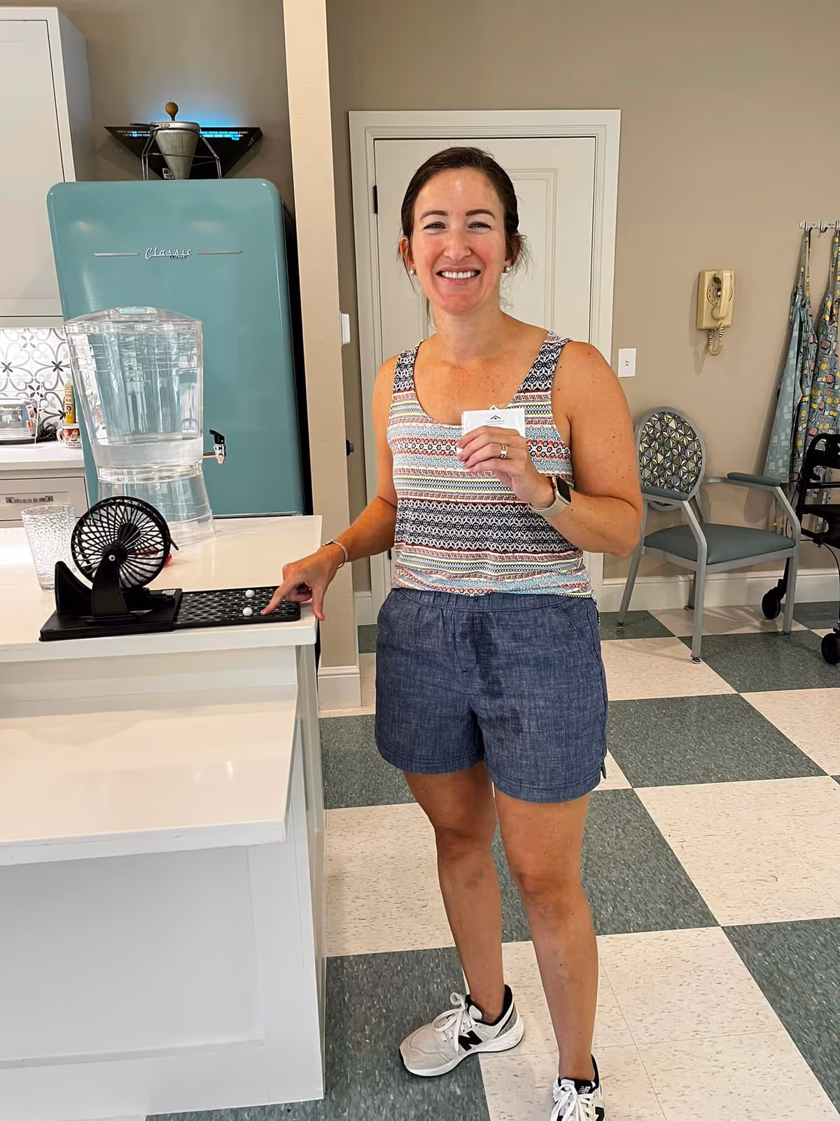 A smiling woman wearing a patterned sleeveless top and blue shorts stands in a kitchen area. She is holding a card in one hand and pressing a button on a small black device with the other hand. Behind her is a light blue retro-style refrigerator, a water dispenser on the counter, a chair, and a wall-mounted telephone.