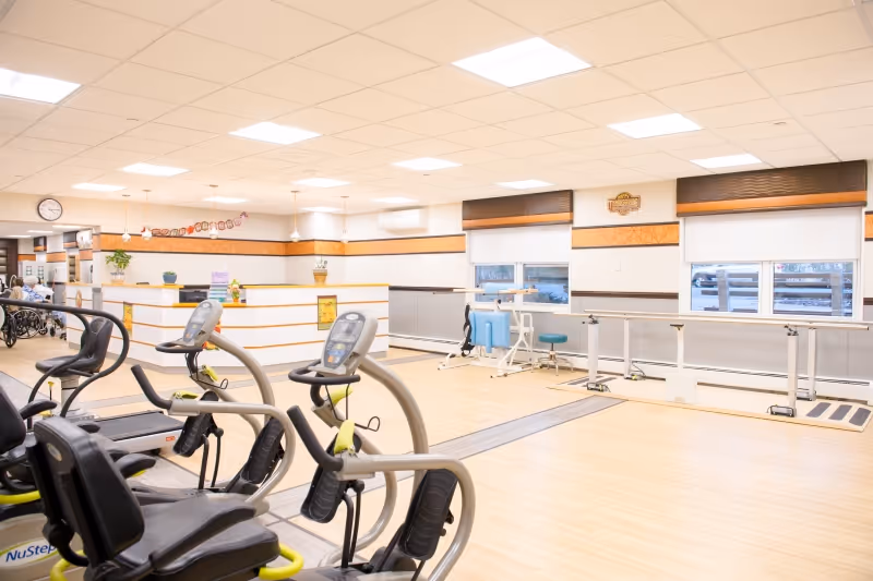 Bright and spacious rehabilitation room with exercise equipment including stationary bikes and parallel bars for physical therapy. The room has light wood flooring, large windows with blinds, and a reception desk area in the background.