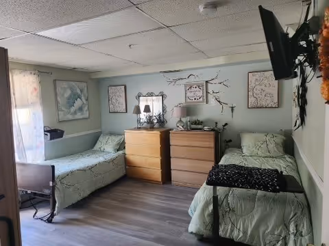A small, tidy bedroom with two single beds on opposite sides of the room, each with light green bedding. Between the beds are two wooden dressers with lamps and decorative items on top. The walls are light green and decorated with framed artwork and a decorative branch. A flat-screen TV is mounted on the wall opposite one of the beds. The floor is wood laminate, and a window with sheer curtains allows natural light into the room.