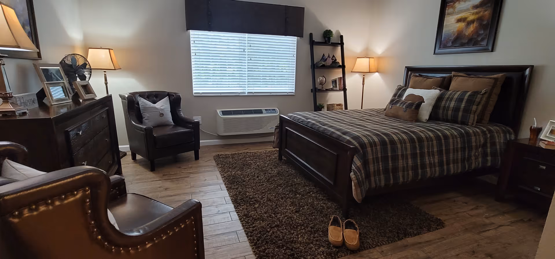A cozy bedroom with a large wooden bed dressed in plaid bedding and multiple pillows. There are two leather armchairs with pillows, a dark wooden dresser with framed photos and a fan, a window with blinds and a dark valance, a brown rug on the wooden floor, a small shelf with decorative items, and two lamps providing warm lighting.