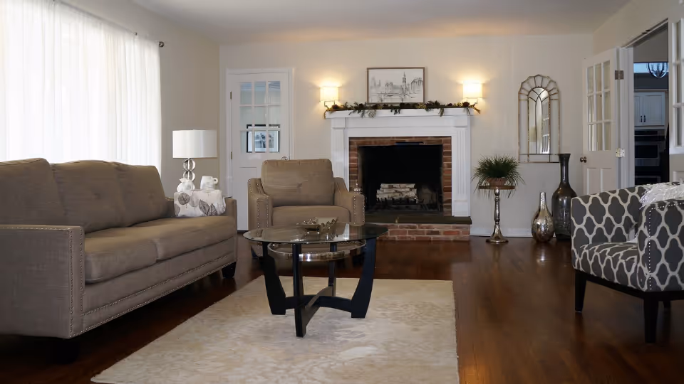 A spacious living room with sofas and armchairs arranged around a glass coffee table facing a brick fireplace with a decorated mantel.