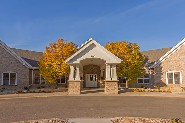 Front exterior view of a single-story brick building with a covered entrance supported by columns. Two trees with yellow autumn leaves flank the entrance. The building number 300 is displayed above the entrance.