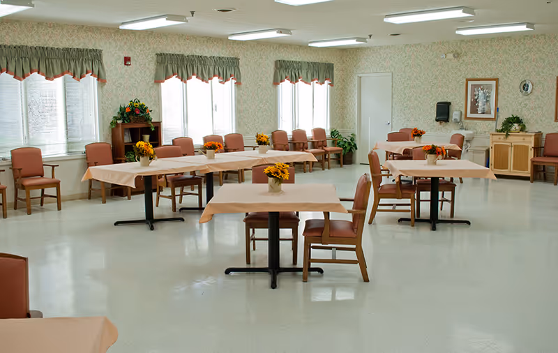 A spacious dining room with several tables covered with beige tablecloths, each adorned with a small vase of yellow flowers. The room has multiple windows with green valances, floral wallpaper, and chairs arranged around the tables and along the walls. There is a wooden cabinet and framed artwork on the walls.