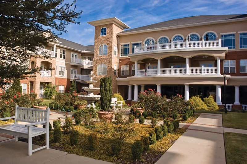 Outdoor courtyard area of a senior living facility with a three-story building in the background featuring balconies and arched windows. The courtyard includes a white bench, a multi-tiered fountain, manicured bushes, and flowering plants under a blue sky.