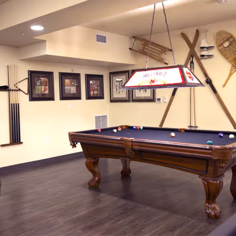 A recreational room with a wooden pool table featuring a dark felt surface and several billiard balls arranged on it. Above the table hangs a rectangular light fixture with a stained glass design. The walls are decorated with framed pictures, a set of pool cues, vintage skis, snowshoes, and a sled. The room has wooden flooring and a warm, inviting atmosphere.