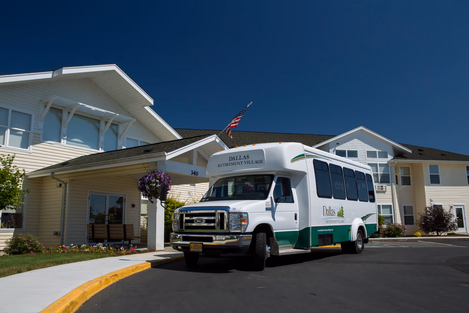 A white and green shuttle bus with the text 'Dallas Retirement Village' parked in front of a light-colored two-story building with a covered entrance and an American flag flying above. The building has multiple windows and is surrounded by landscaping with flowers and bushes under a clear blue sky.