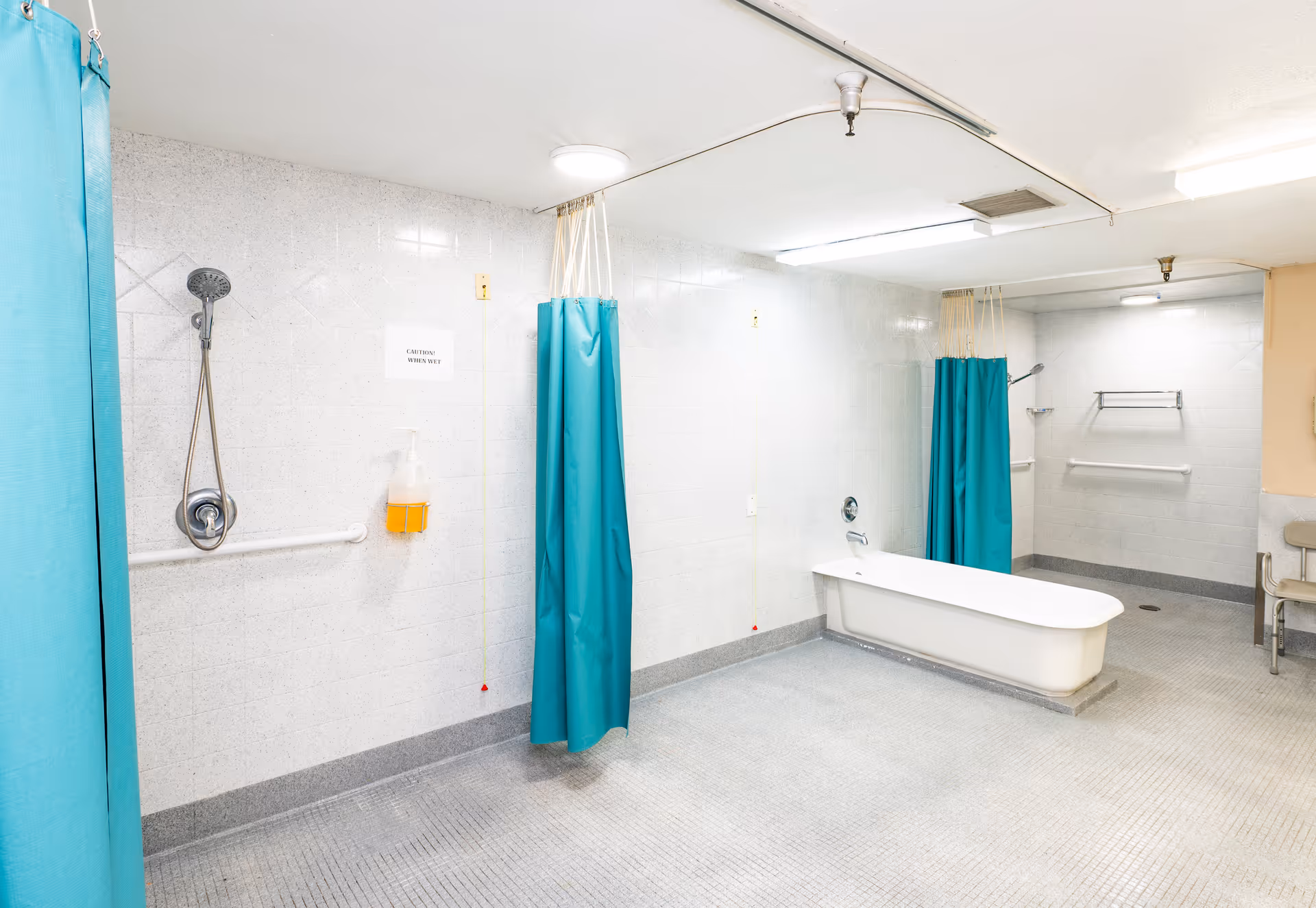 A spacious, institutional-style bathroom with white tiled walls and gray tiled floor. The room features a white bathtub, two shower areas with teal privacy curtains, a handheld showerhead, grab bars, a soap dispenser, and a chair in the corner. The lighting is bright with ceiling fixtures.