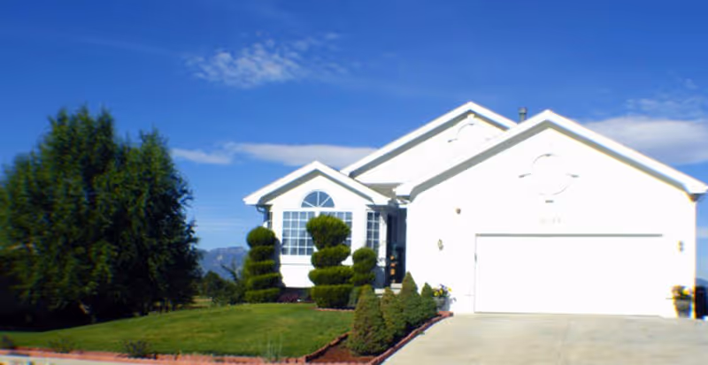 A white single-story house with a two-car garage, manicured lawn, and sculpted bushes under a blue sky with some clouds.