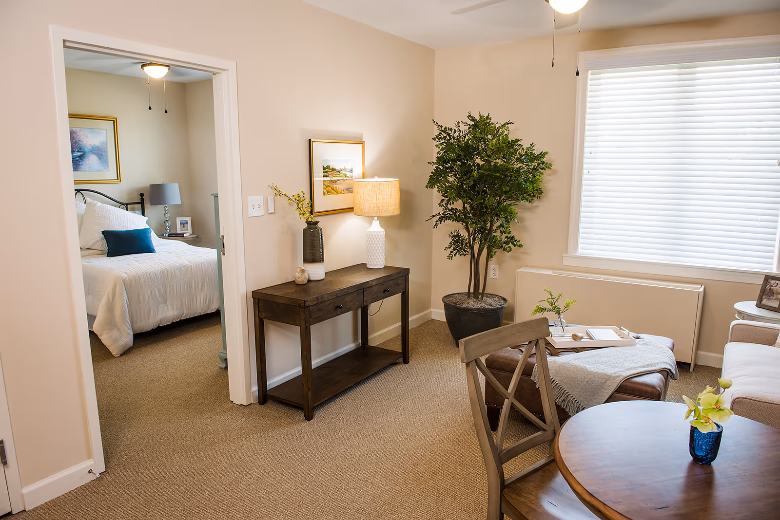 A cozy living room area with a wooden table and chair in the foreground, a beige sofa, a large potted plant, and a wooden console table with a lamp and decorative items. Through an open doorway, a bedroom with a bed, pillows, a nightstand, and a framed picture on the wall is visible.