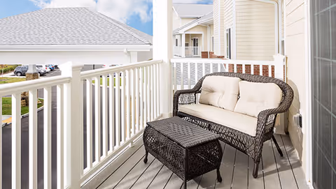 A small outdoor balcony with a cushioned wicker loveseat and matching wicker coffee table. The balcony has white railings and overlooks a parking area and neighboring buildings under a partly cloudy sky.